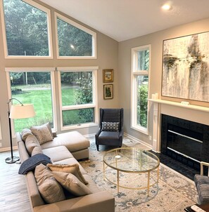 Un salon comprenant un canapé, un fauteuil et une cheminée. De grandes fenêtres offrent une vue sur l’extérieur.