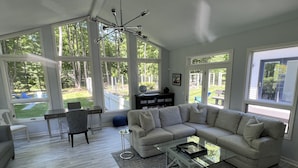 A spacious living room with a large sectional sofa, a glass coffee table, and a view of the backyard through large windows. - Ultimate Family Retreat: Pool, Huge Yard, Gardens, Chickens, 5 Beaches Closeby (Fennville)