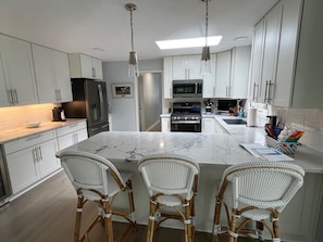 A modern kitchen with white cabinets, a marble island, wicker chairs, and stainless steel appliances.