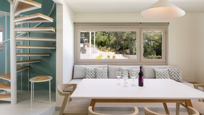 A modern kitchen with a white table, wooden chairs, a built-in bench, and a view of the outdoors through large windows.