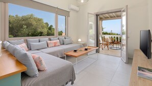 A modern living room with a sofa, a coffee table, and a TV. There is a balcony with a table and chairs.