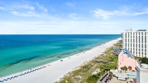 A beach with a white sandy shore, clear turquoise water, and a hotel with a pink building and a white facade. - Penthouse Perfection! Simple Elegance at Tops'l Beach Manor 1408 w/ Breathtaking Gulf Views (Miramar Beach)