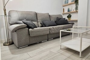 A grey sofa with multiple cushions, a glass coffee table, a floor lamp, and shelves with plants.