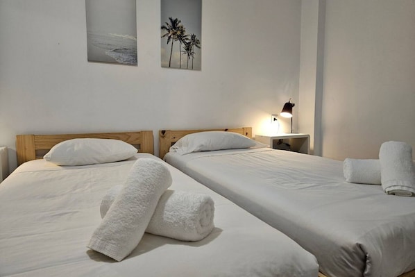 A hotel room with two beds, white linens, a nightstand with a lamp, and wall art depicting palm trees and a beach.