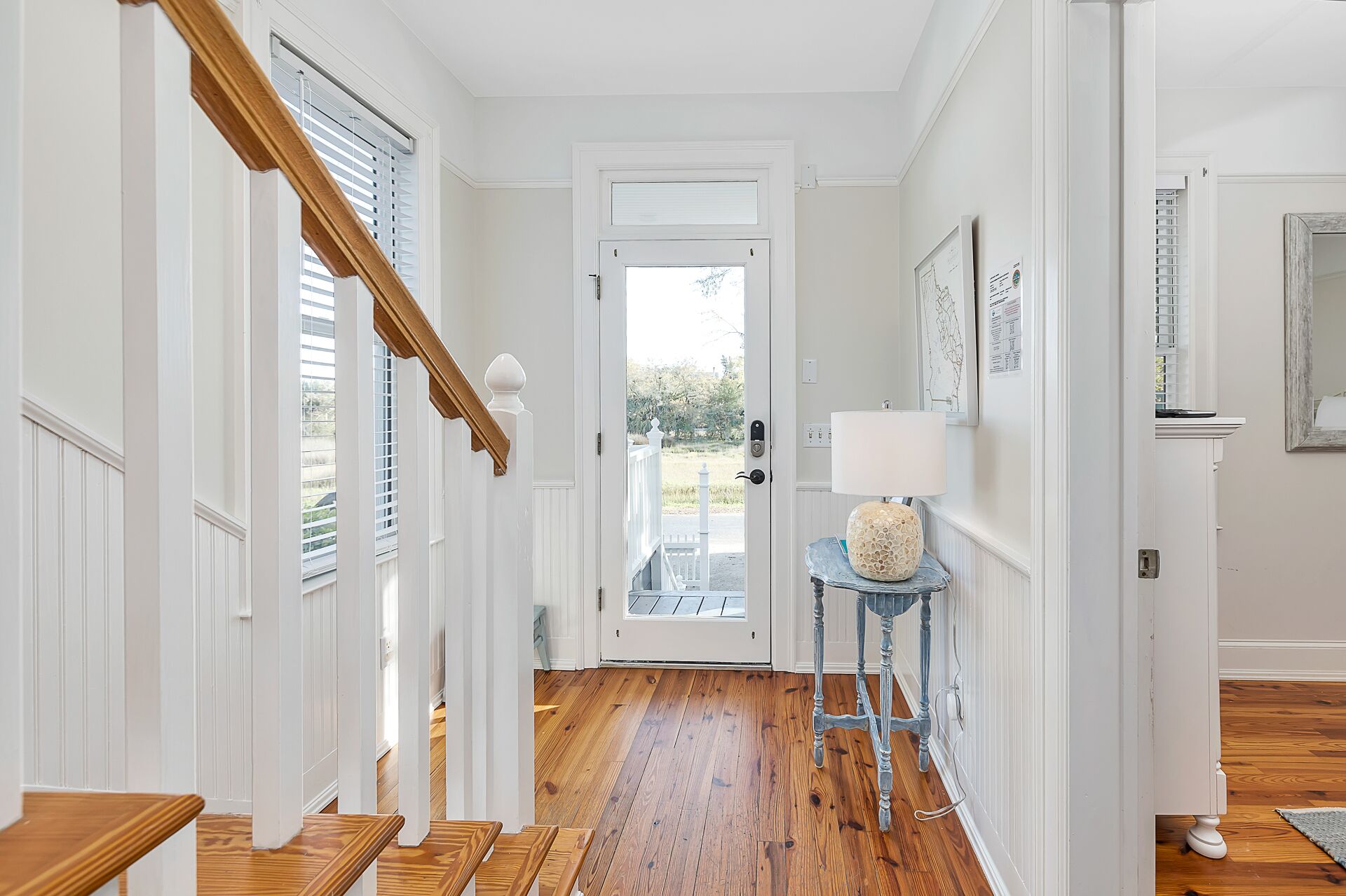 Un couloir avec un escalier en bois, des murs blancs et une porte donnant sur l’extérieur.