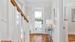 Un couloir avec un escalier en bois, des murs blancs et une porte donnant sur l’extérieur.