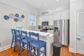 A modern kitchen with white cabinets, a stainless steel refrigerator, a microwave, and a sink. There are blue chairs around a small dining table.