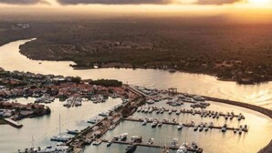 An aerial view of a coastal marina with numerous boats docked, surrounded by a town with visible buildings and a river. - Chic luxury condo at Los Altos Casa de Campo (La Romana)