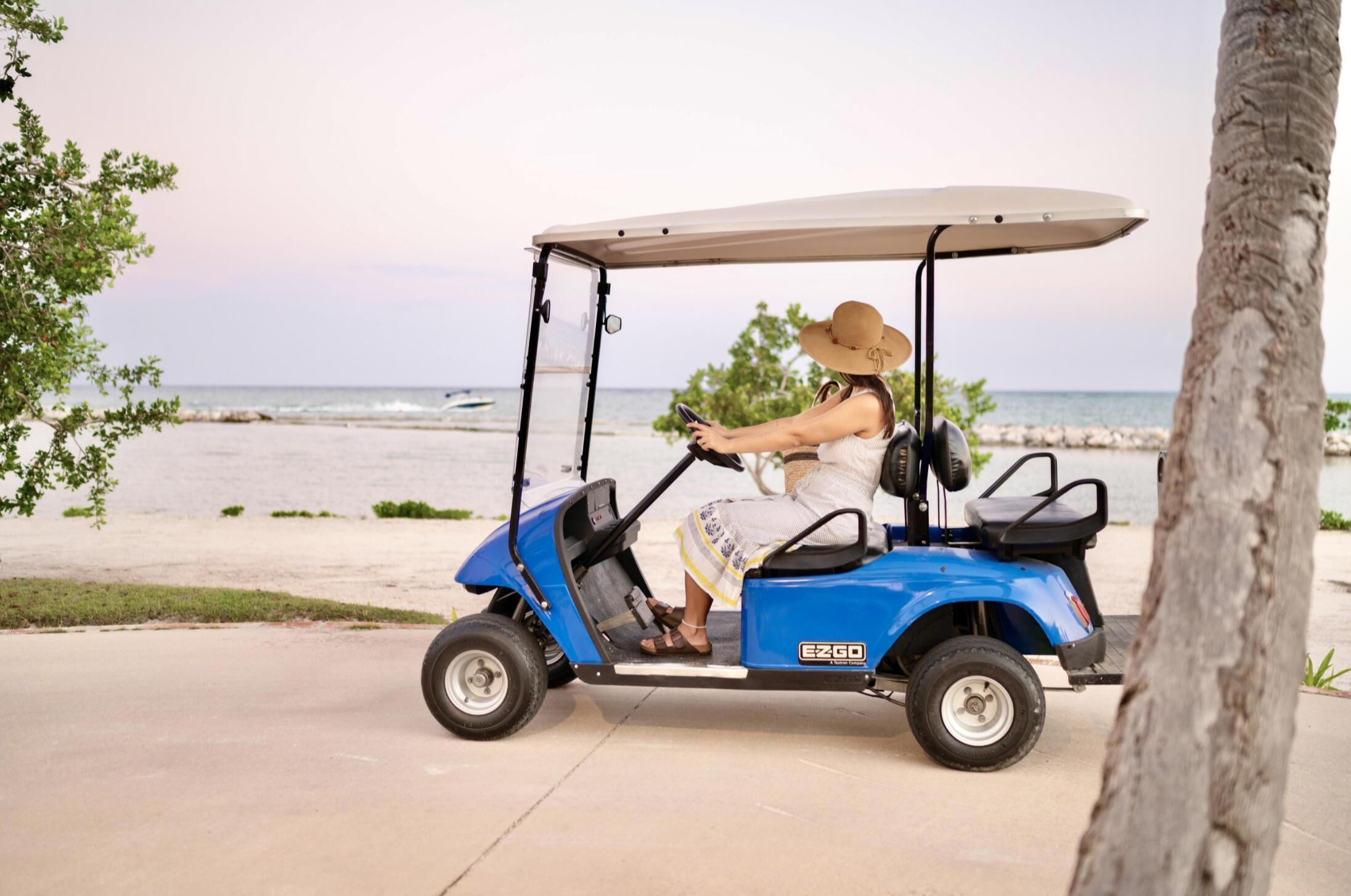 Una persona conduce un carro de golf azul con toldo por un camino pavimentado cerca de la playa.