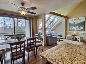 A spacious living area with a dining table, chairs, a sofa, and a large window. - SunBear- AC, Pool, Game Room, Fireplace, Hiking! (Killington)