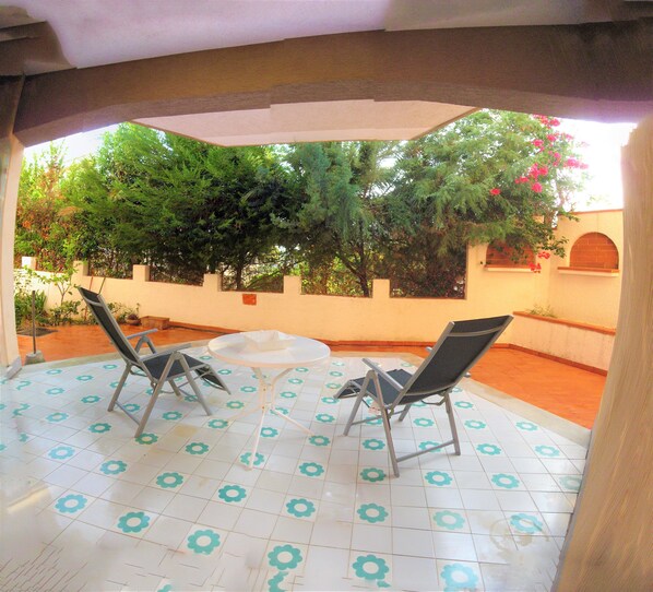 Un patio al aire libre con dos sillas y una mesa, rodeado por un muro blanco y un jardín con árboles y flores.