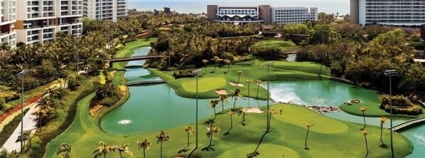 Ein Golfplatz mit einem Wasserhindernis, Palmen und modernen Gebäuden im Hintergrund.