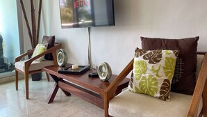 Een woonkamer met een flatscreen televisie aan de muur, een houten salontafel en twee fauteuils met kussens.