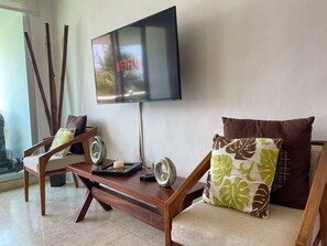 Un salon avec un téléviseur à écran plat fixé au mur, une table basse en bois et deux fauteuils avec des coussins.