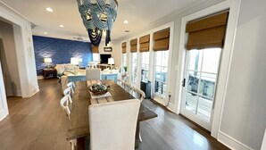 A spacious living room with a large dining table, a blue accent wall, and a view of the outdoors through large windows.