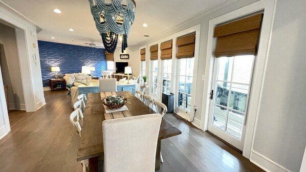 A spacious living room with a large dining table, a blue accent wall, and a view of the outdoors through large windows.