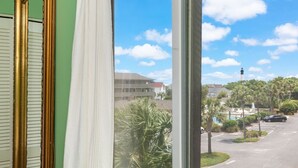 A view from a window with a curtain, overlooking a street with palm trees and buildings. - Lighthouse Point 13B | Oceanfront Condo, Pool! (Tybee Island)