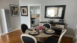 A dining area with a round table set for four, surrounded by four chairs with dark cushions. There is a small vase with pink flowers on the table. - The Heart of Belmont Shore (Long Beach)