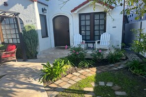 Une maison à l’extérieur blanc, avec une porte marron foncé et un petit patio comprenant une chaise et une table, toutes deux blanches.