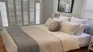 A bedroom with a bed, a nightstand, a lamp, a framed picture on the wall, and a window with blinds. - The Heart of Belmont Shore (Long Beach)