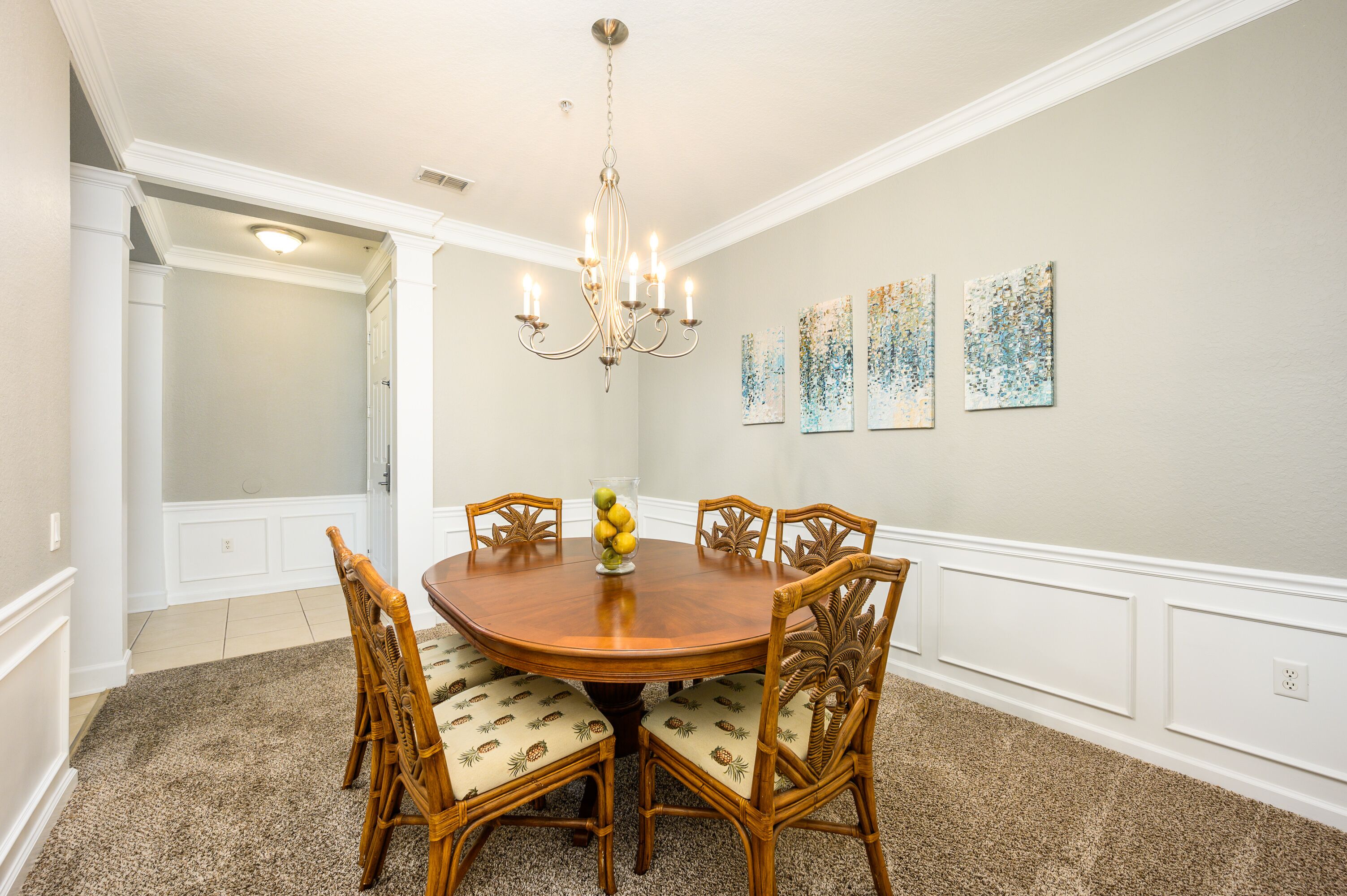 A dining room with a round wooden table, six chairs, a chandelier, and four abstract paintings on the wall.