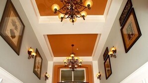 Ein Flur mit hoher Decke, einem Chandelier und einem Balkon. Zwei Holztische mit Stühlen und Blumentöpfen sind vorhanden.