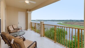 Ein Balkon mit Glastisch und Stühlen, einem Deckenventilator und Blick auf einen Fluss sowie entfernte Gebäude.