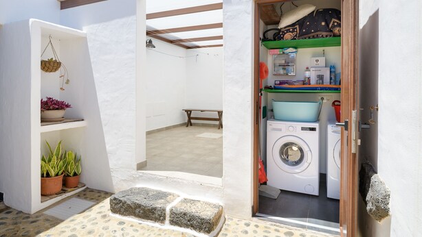 Un espacio para lavandería con lavadora, lavabo y tendedero. Hay plantas en macetas y un banco en el área exterior contigua.