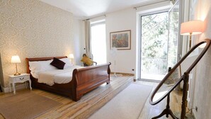 Een slaapkamer met een houten bed, nachtlampjes, een kamerkleed met een patroon en een groot raam met uitzicht op bomen.
