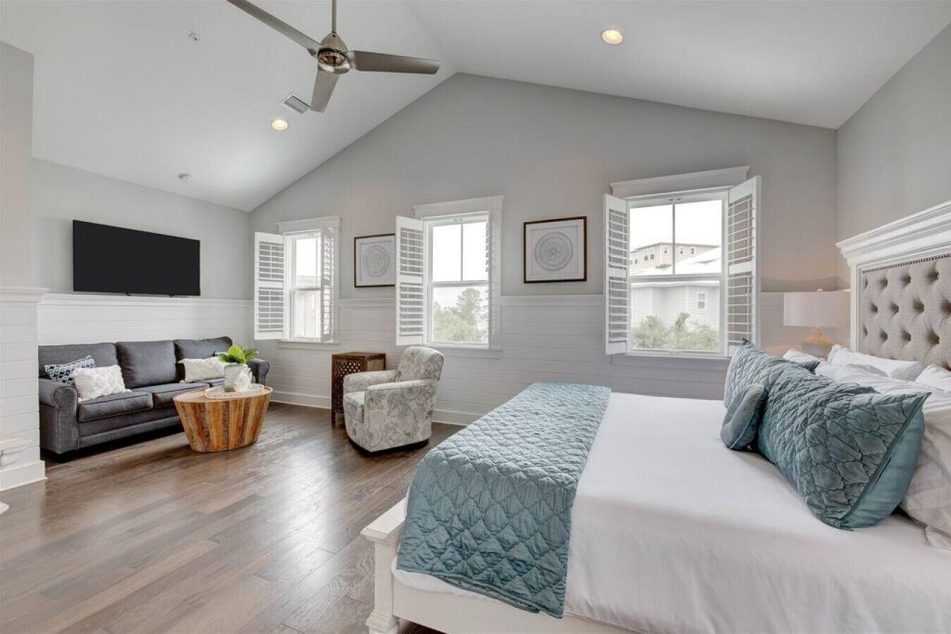 A bedroom with a large bed, a gray sofa, a chair, and a TV on the wall.