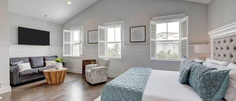 A bedroom with a large bed, a gray sofa, a chair, and a TV on the wall.