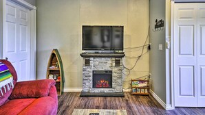 A living room with a stone fireplace, a flat-screen TV mounted above it, a red sofa, a bookshelf, and a door. - Hidden Gem: Dock & Views on Pine Hollow Reservoir! (Tygh Valley)