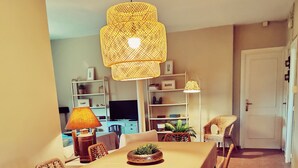 A dining area with a long table, wicker chairs, a hanging light fixture, and a television in the background.