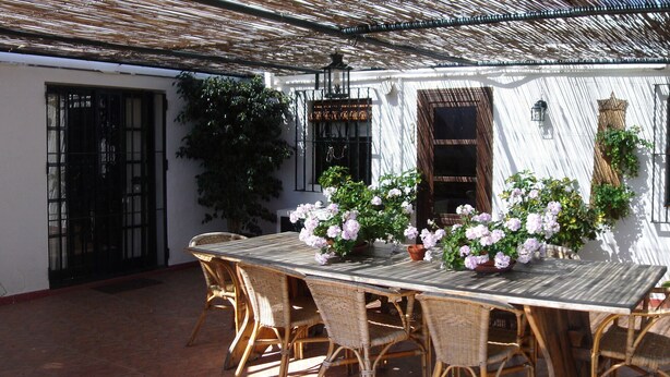 A patio with a table and chairs, surrounded by potted plants and a white wall.