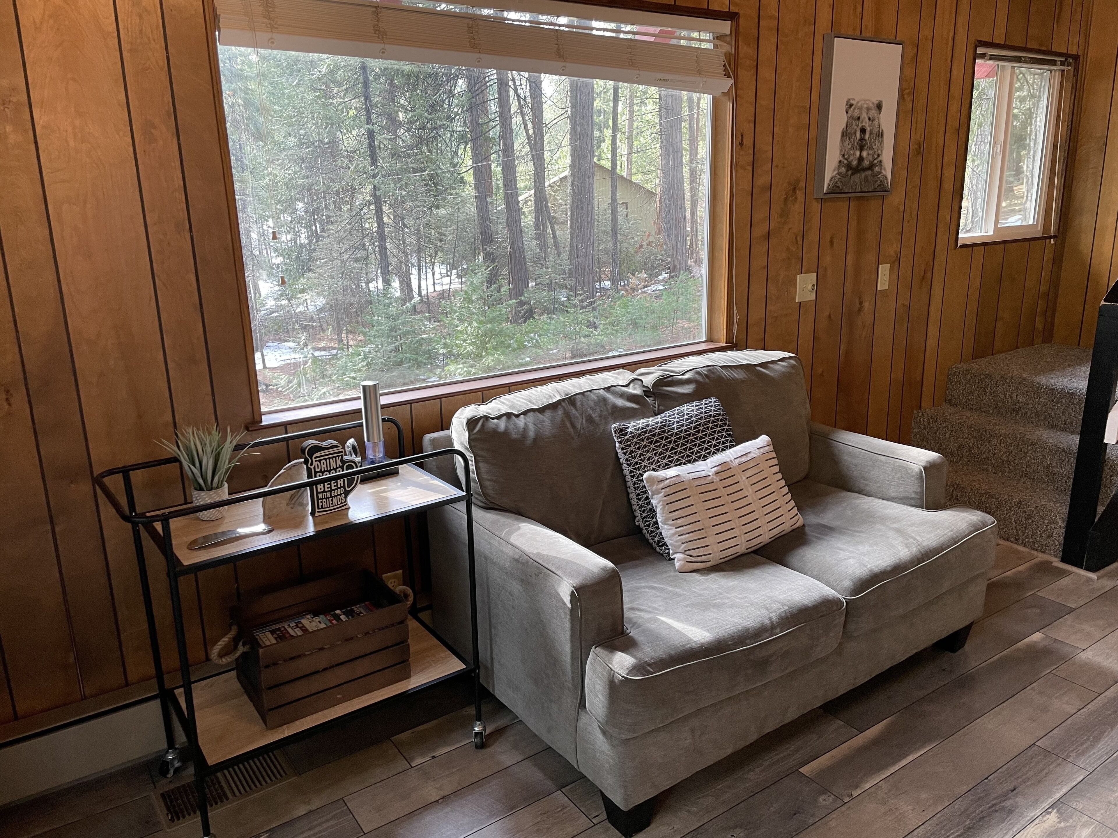 A cozy room with a large window, a grey sofa, a small table with a plant, and a framed picture on the wall.