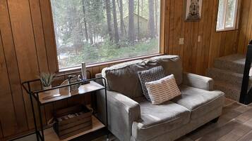 A cozy room with a large window, a grey sofa, a small table with a plant, and a framed picture on the wall.