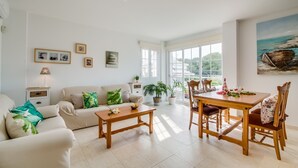 A spacious living area with a sofa, a wooden coffee table, and a dining table with chairs. There are potted plants and artwork on the walls.