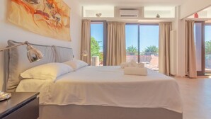 Ein großes Bett mit weißer Bettwäsche, eine Nachttischlampe und Blick ins Freie durch große Fenster.