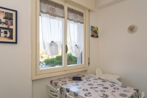 Een kamer met een tafel en stoelen, een raam met gordijnen, een klok aan de muur en een ingelijste foto.