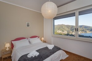Een slaapkamer met een bed, een nachtkastje, een lamp en een raam met uitzicht op een landschap.
