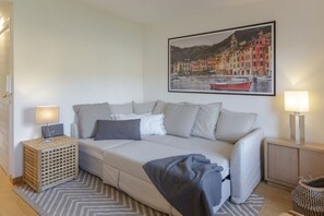 A modern bedroom with a sofa bed, a small bedside table, a lamp, a framed picture on the wall, and a patterned rug.