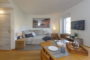 A cozy living room with a sofa, a dining table set with plates and cups, a television, and a painting of a cityscape.