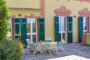 Un patio al aire libre con una mesa y sillas, una pequeña área ajardinada y un edificio con contraventanas verdes y detalles decorativos.