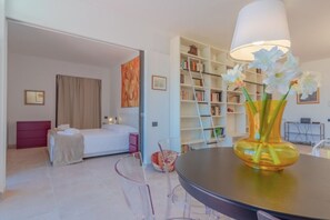 Una habitación de hotel moderna con una cama, un escritorio con lámpara, una estantería y una mesa de vidrio transparente.