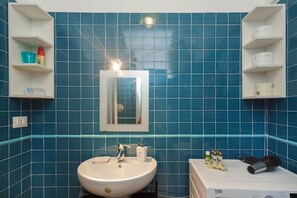 Un baño con azulejos azules, un lavamanos blanco, un espejo y estantes con artículos de aseo.