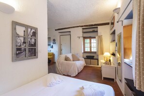 Una habitación con una cama, un sofá tapizado, una mesita de noche con una lámpara, un cuadro enmarcado en la pared y una ventana con cortinas.