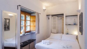 Un dormitorio con cama, ventana con cortinas, mesita de noche, espejo y estante con una botella.