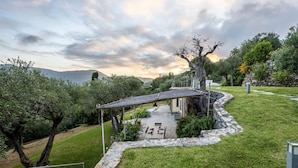 Une maison avec un chemin de pierre menant à un espace extérieur aménagé et abrité, entouré de verdure et offrant une vue sur des collines lointaines.