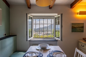 A dining area with a table set for two, a window with an open shutter, and a view of a mountainous landscape.