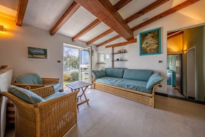 A room with a wicker sofa, two wicker chairs, a small table, wooden beams on the ceiling, and a view through a glass door.
